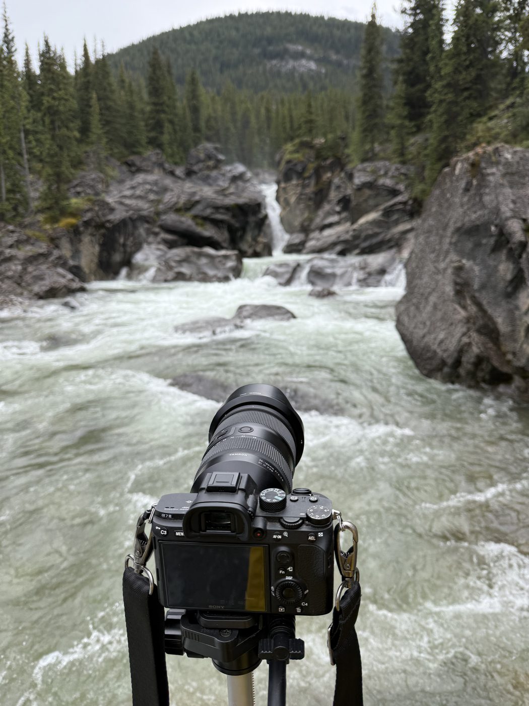 The back of a Sony mirrorless camera with a Tamron zoom lens on a tripod, framing a waterfall in a rocky canyon