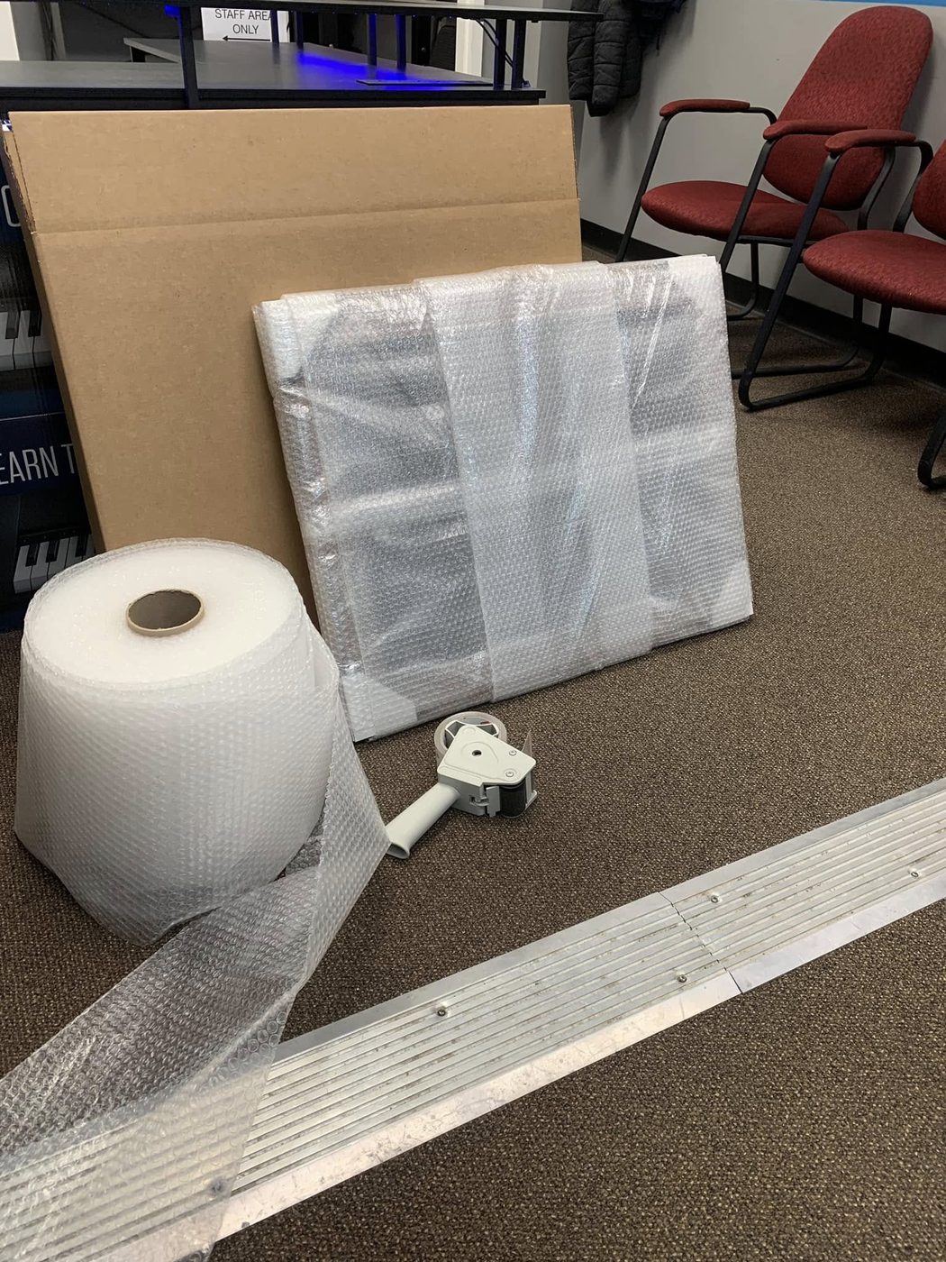 A framed print wrapped in bubble wrap standing in front of its cardboard box, with a bubble wrap roll and tape gun in front