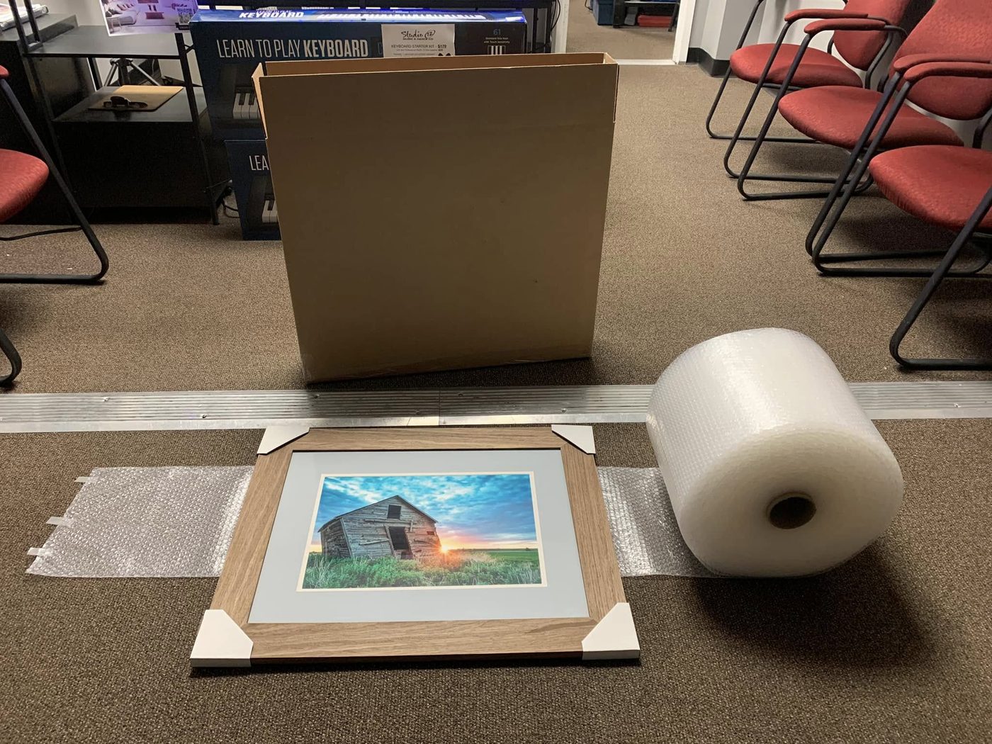 A matted and framed print of a weathered prairie barn at sunset laid out beside a cardboard shipping box and a roll of bubble wrap